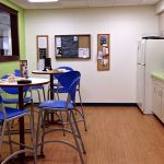 After: This workplace kitchen was enlarged during an office renovation.  Bright colors, high-top tables, a wood-look vinyl floor, and windows to the hall make this space welcoming.  Employees now use this break room for eating, socializing, and collaboratin 